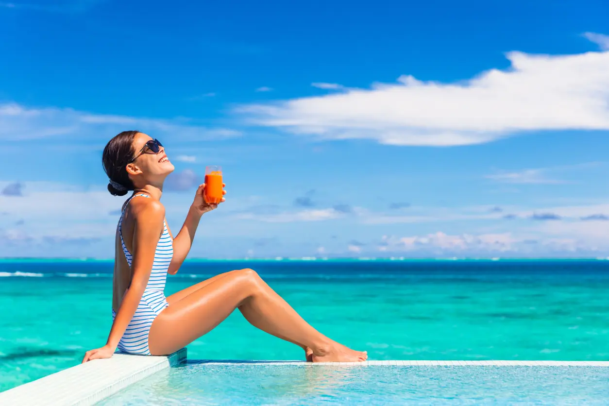 Woman in a striped swimsuit sits at the edge of a pool, holding a glass of orange juice, with the ocean and blue sky in the background.