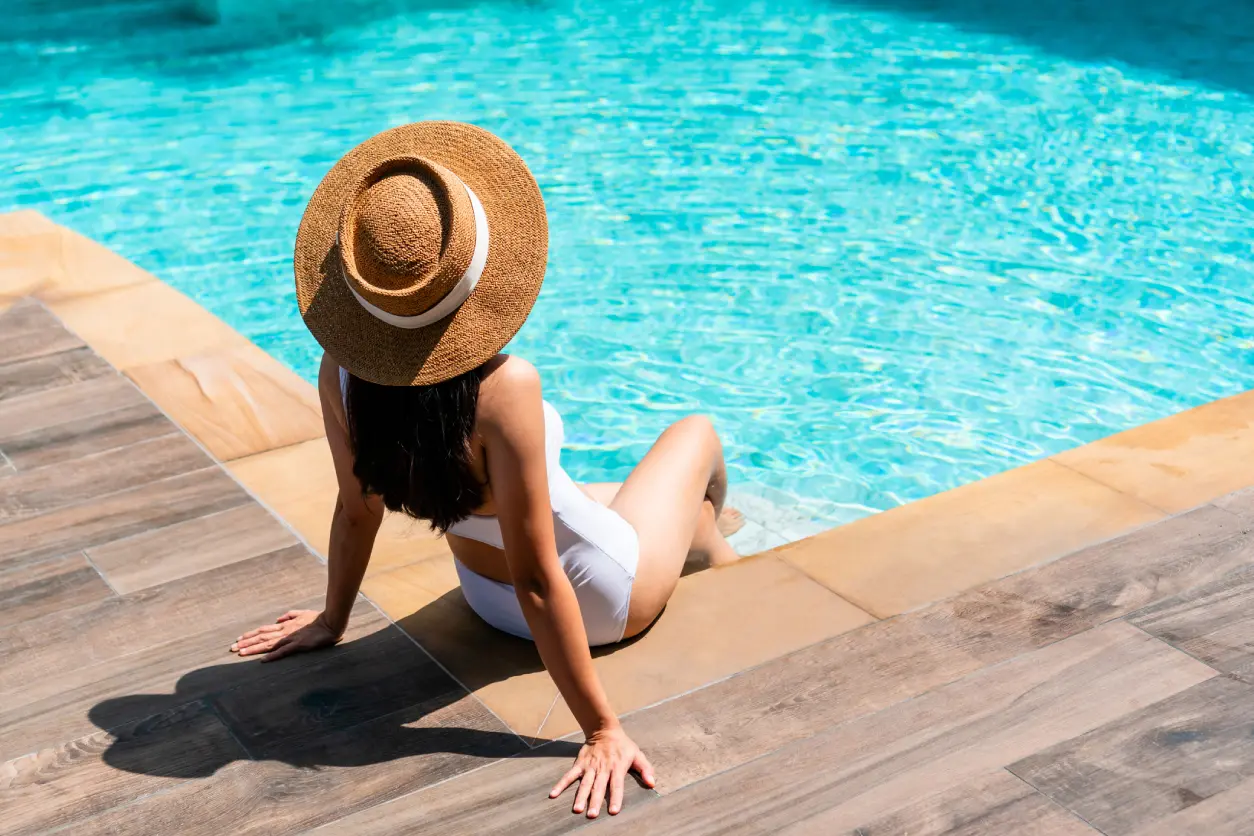 Woman in a white swimsuit and straw hat sitting by the edge of a pool, with her feet in the water and hands resting on wooden decking.